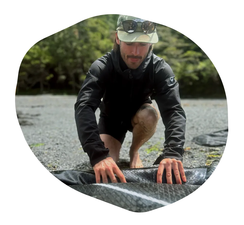 A young man kneeling, rolling up a rubber mat on a rocky beach.